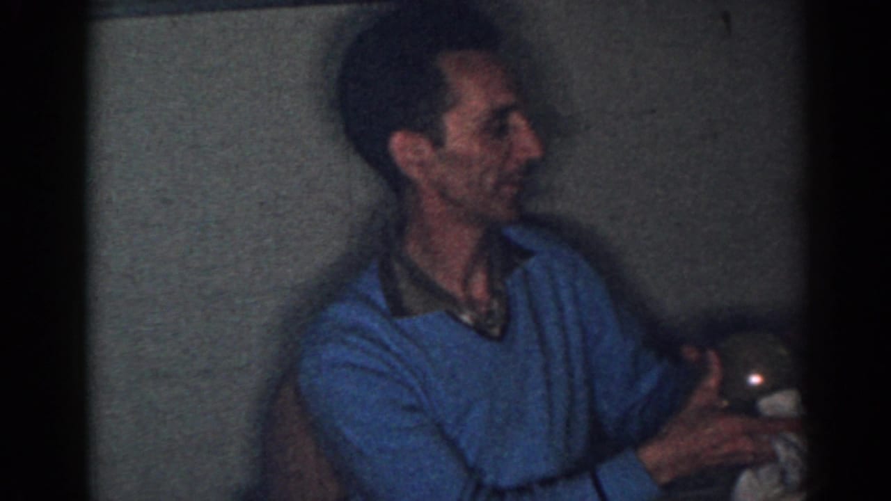 Man in a blue sweater sits indoors holding a white cup in dim light.