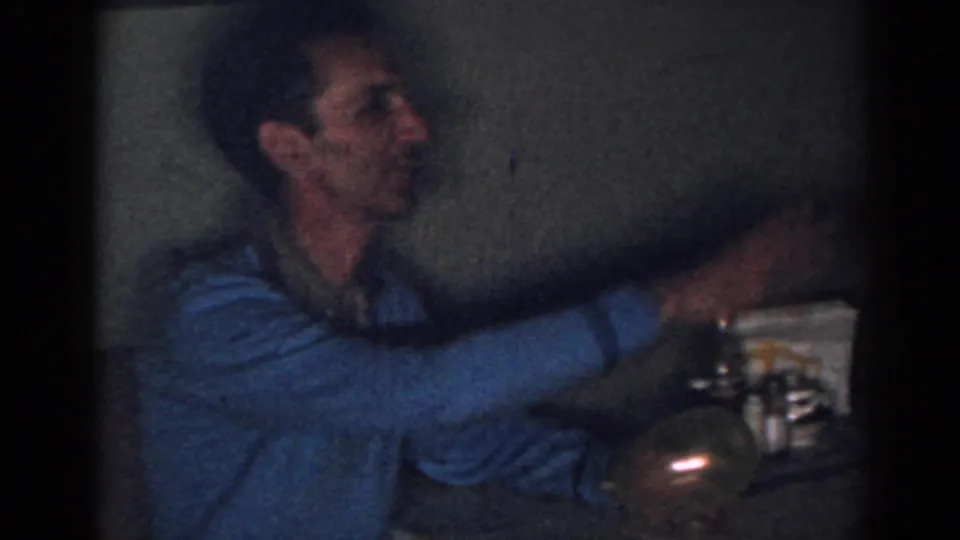 A man in a blue sweater reaches across a dim New Jersey interior toward cups or tableware in the 1963 footage.