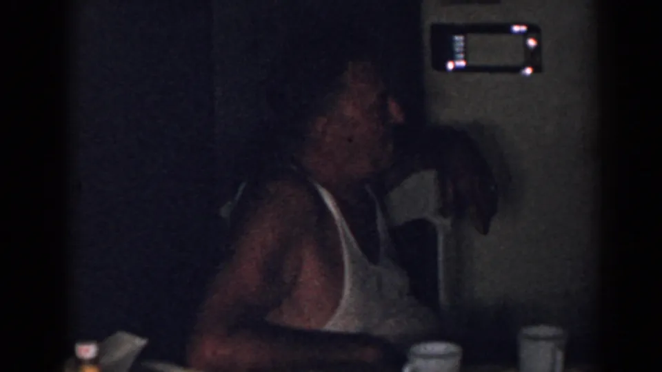 A man in a white tank top sits at a table with mugs in a dim private interior, recorded as 1963 New Jersey footage.