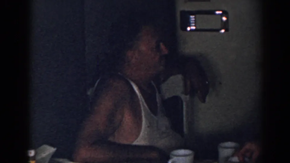 The seated man in a white tank top remains by the table as another person's hand appears at the edge of the dim room.