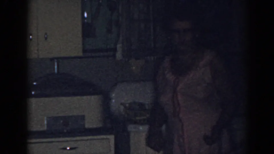 1963 New Jersey home-movie frame of a woman in a light apron standing in a dim kitchen beside cabinets and counter space.