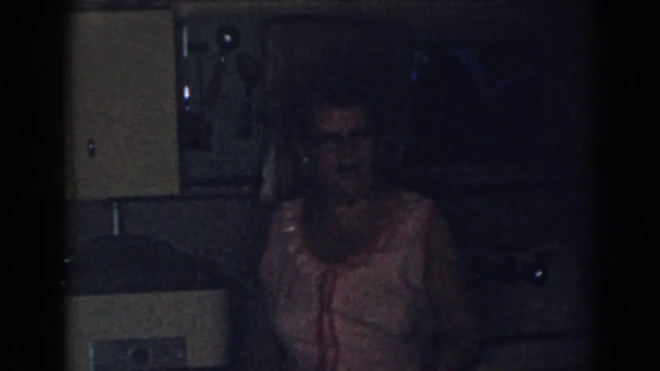 1963 New Jersey kitchen frame showing a woman in a light apron facing the camera amid cabinets, counter surfaces, and deep shadows.