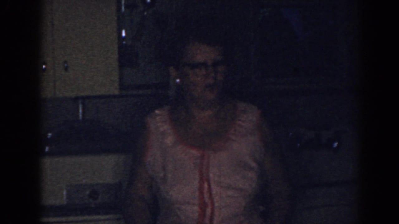 Adult woman in a pink sleeveless dress stands in a dim kitchen.