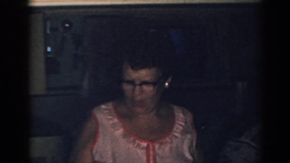 The woman in glasses and a pink dress appears near kitchen cabinetry in this dim 1963 New Jersey home-movie frame.