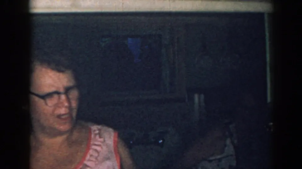 A woman in a pink dress and glasses is partially framed in a dark kitchen during the 1963 New Jersey home movie.