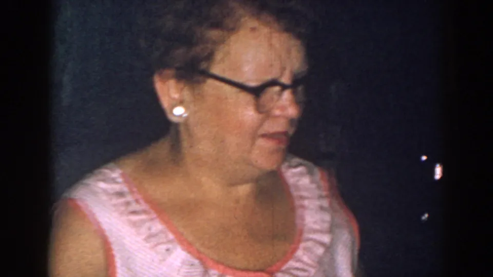 The woman, wearing glasses and pearl earrings, turns in a dim New Jersey kitchen in this 1963 8mm home-movie frame.