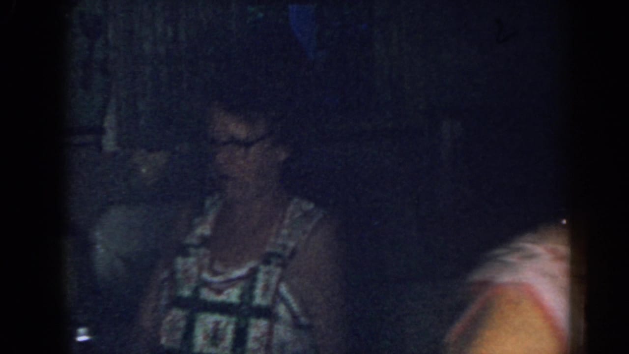 Older woman in glasses stands in a dim kitchen beside cabinets and a counter.