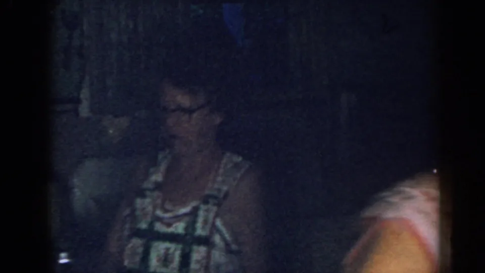 Older woman in glasses and a patterned sleeveless dress stands in a dim 1963 New Jersey kitchen interior.