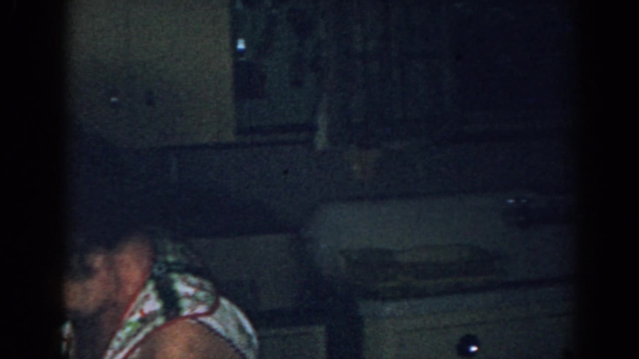 Older woman bends forward near a kitchen counter while cabinets and a sink area sit behind her.