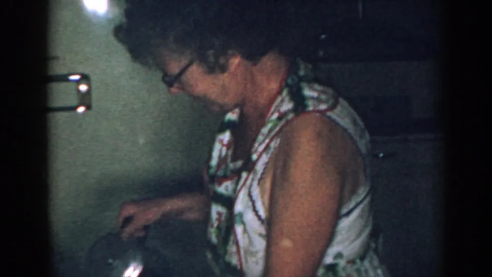 Older woman in glasses reaches toward a kettle or pot at the counter in a dim 1963 New Jersey kitchen.