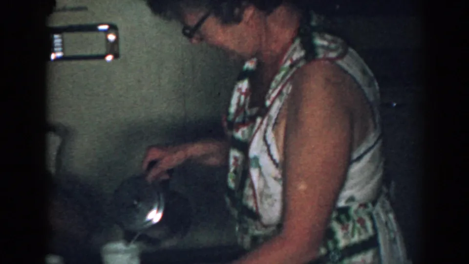 In a 1963 New Jersey home-movie frame, a woman in a patterned sleeveless dress pours coffee near white mugs in a kitchen.