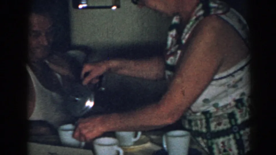A seated man in a white tank top watches a woman pour coffee at a kitchen table in a 1963 New Jersey home interior.