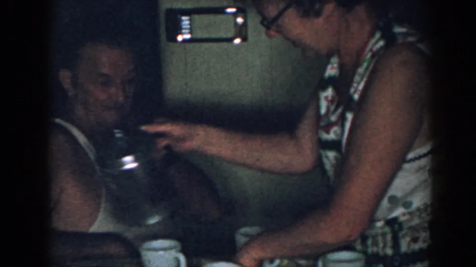 A woman in glasses pours coffee from a glass pot near a seated man and white mugs in a 1963 New Jersey kitchen scene.
