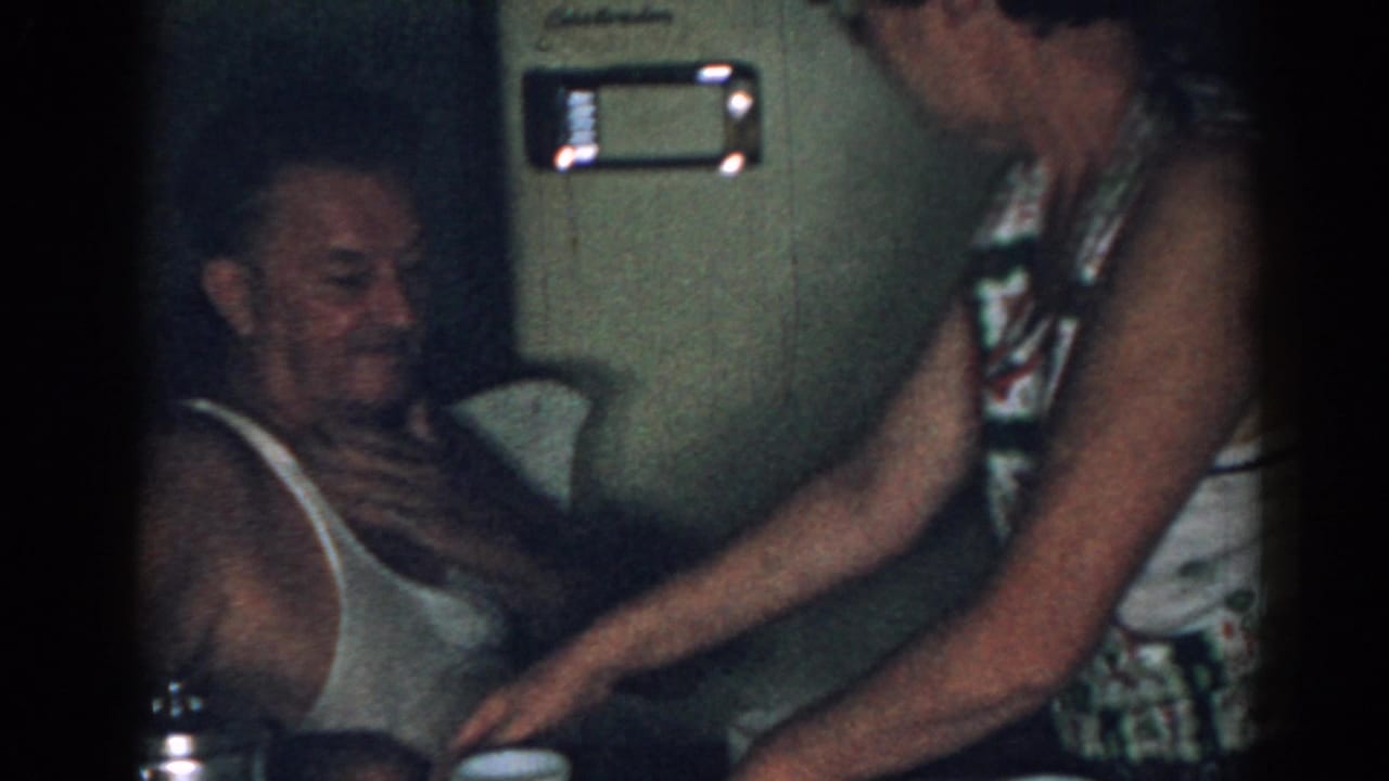 Older man seated in a tank top while a woman stands beside him in a dim room.