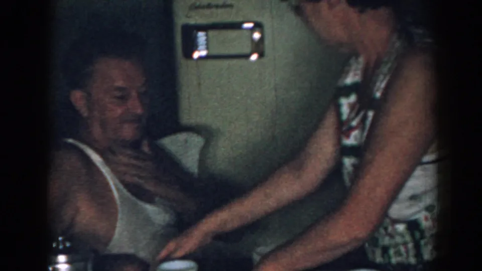 In a 1963 New Jersey home-movie frame, an older man sits in a white tank top as a woman in a patterned sleeveless dress tends to him.