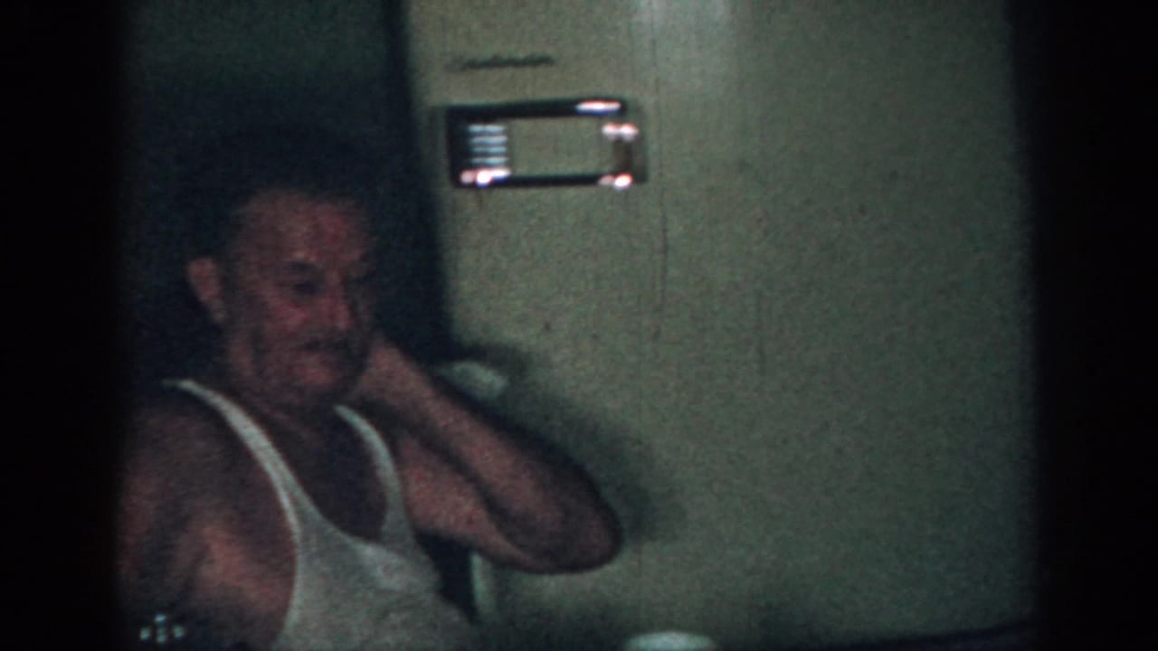 The older man remains seated in the dim New Jersey interior, his hand raised near his neck beside a wall-mounted appliance.