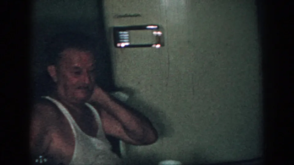 The older man remains seated in the dim New Jersey interior, his hand raised near his neck beside a wall-mounted appliance.