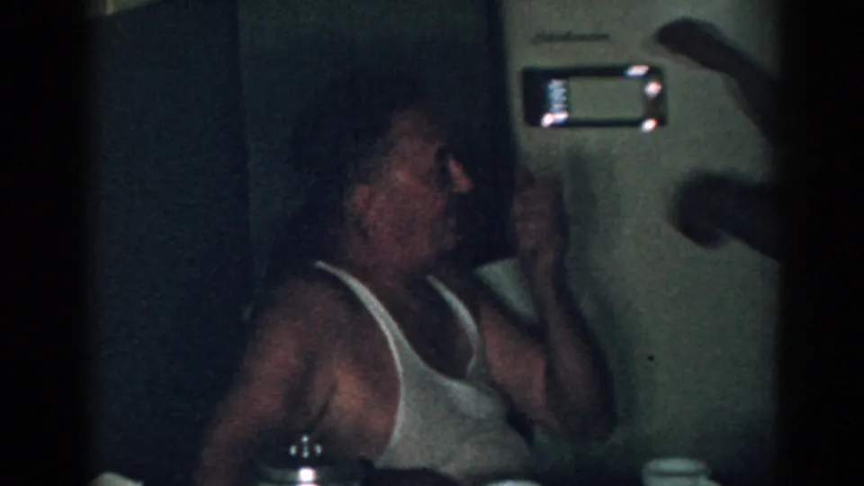 The older man leans back in his chair with one hand raised, suggesting discomfort from heat inside the 1963 New Jersey home.