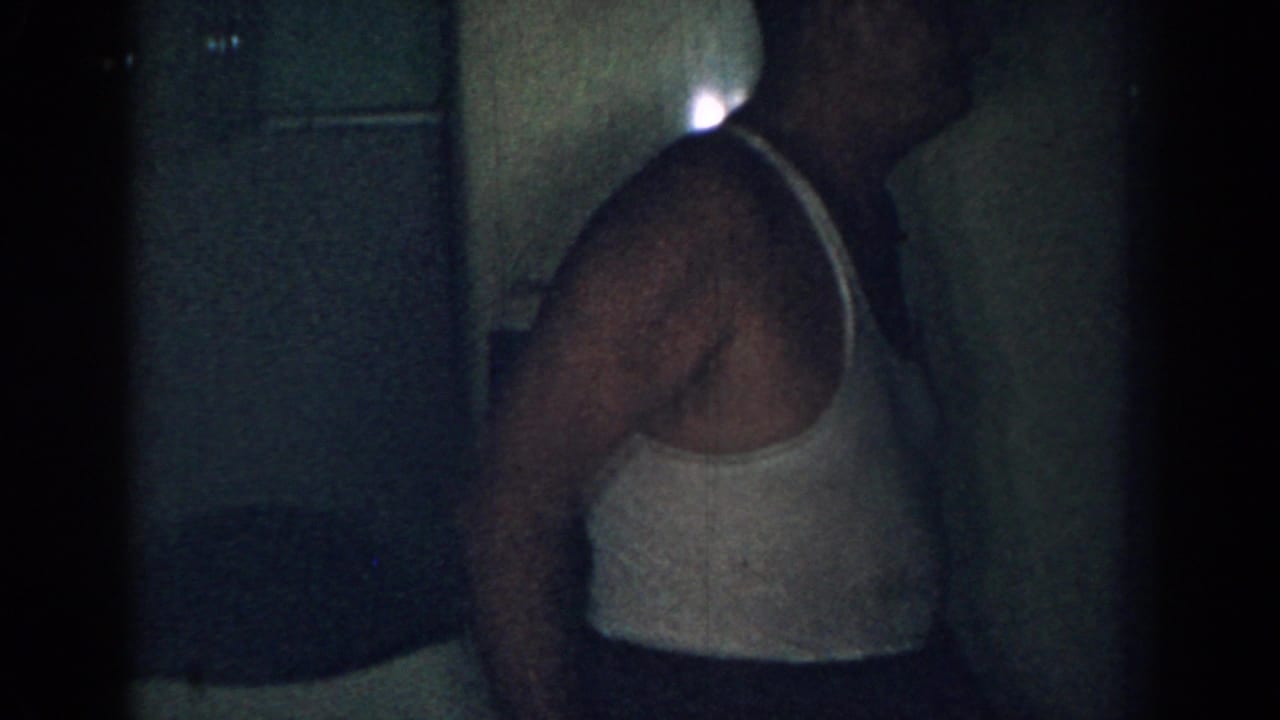 Adult man in a white tank top standing beside a refrigerator in a dark kitchen.