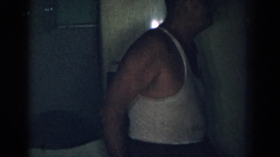 Adult man in a white tank top near a refrigerator in a dim 1963 New Jersey home kitchen.