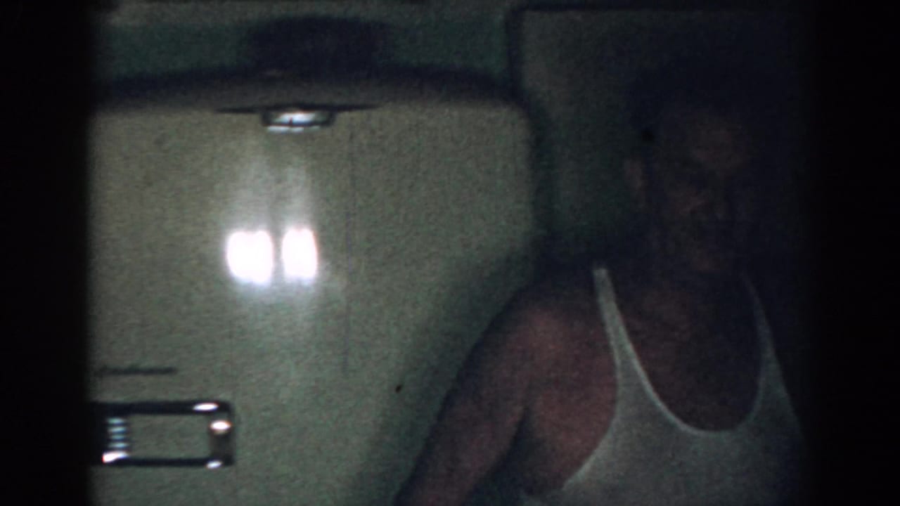 Man in a white tank top partly framed beside pale kitchen cabinets and a refrigerator.