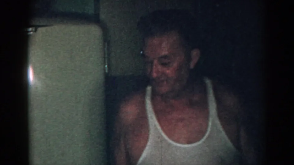 Adult man in a white tank top looks downward near a refrigerator in a grainy 1963 New Jersey kitchen scene.