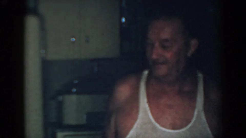 Adult man in a white tank top appears to smile or look down in a dim 1963 New Jersey kitchen interior.