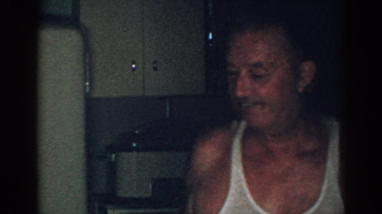 Older man in a white undershirt smiling in a dim interior room.