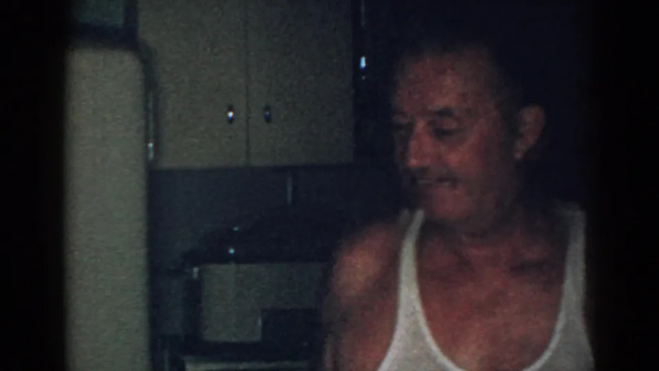 Older man smiles inside a dim New Jersey home interior in 1963, filmed with soft 8mm grain and low light.