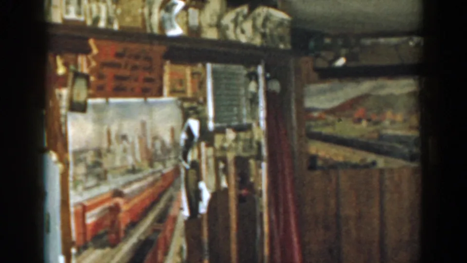 1963 New Jersey home-movie frame with a red train backdrop, pinned wall images, mesh screen, and a red curtain at right.