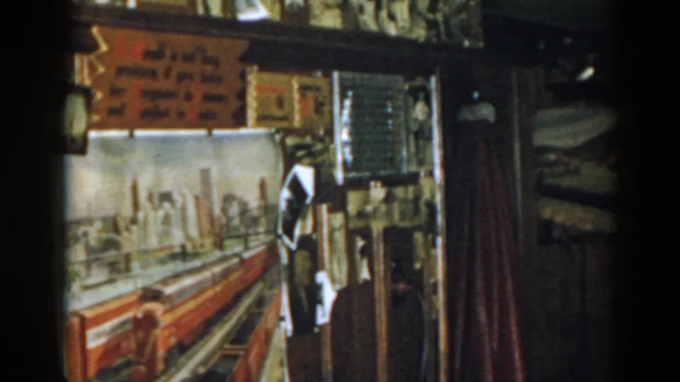 1963 New Jersey 8mm frame of a dim interior with train artwork, red curtain, mesh screen, and wall-mounted pictures.