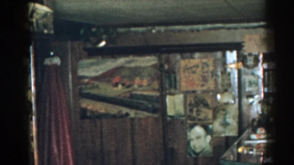 1963 New Jersey home-movie frame of a densely decorated private room with railroad imagery, wall memorabilia, and a partial face or bust visible.