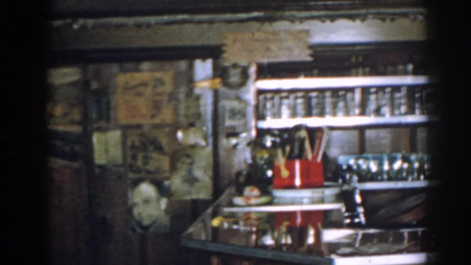 1963 New Jersey home-movie frame of a wood-paneled bar or serving area with glassware, wall decor, and counter objects.