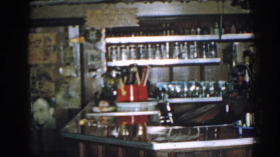 1963 New Jersey home-movie frame of a bar interior with shelves of bottles, glassware, a red holder, and a blue tray.