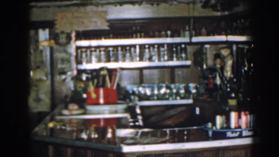 1963 New Jersey 8mm frame showing a dim bar counter with glassware, bottle shelves, and colorful bottles in a blue tray.