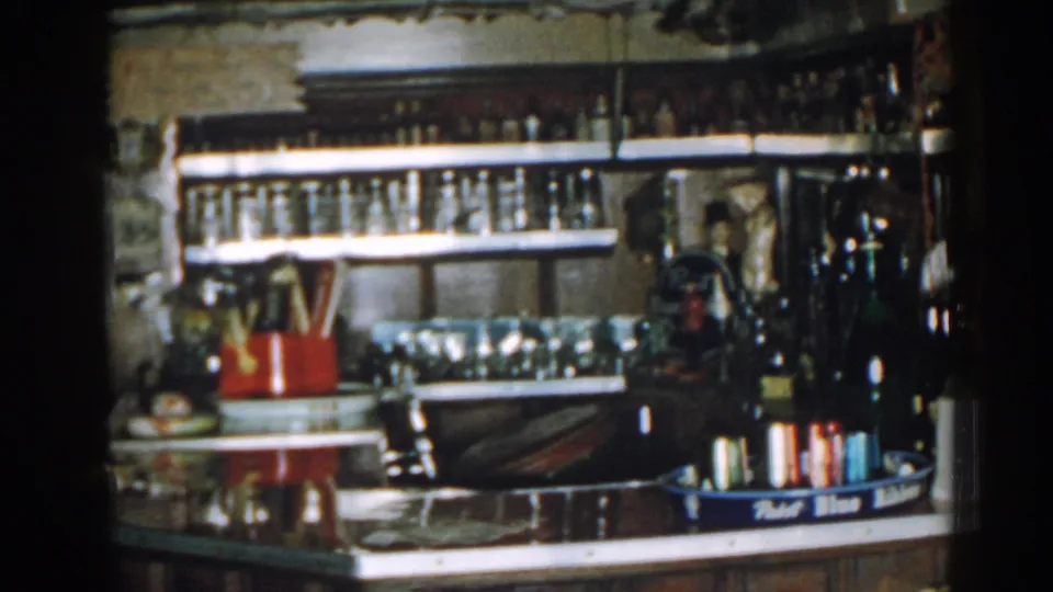 1963 New Jersey home-movie frame of a vintage bar counter with bottle shelves, glassware, a red holder, and trayed bottles.