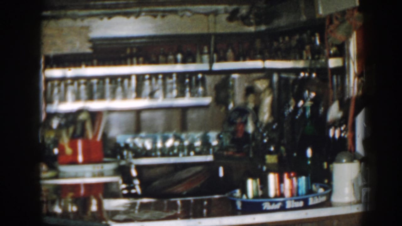Cluttered shop counter with bottle shelves, glassware, a red container, and small cans in a blue tray.