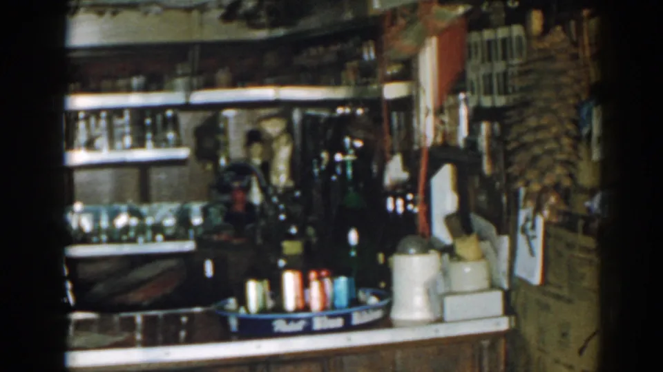 1963 New Jersey 8mm view of a crowded shop counter with bottles, shelving, a red container, and wall-mounted display items.