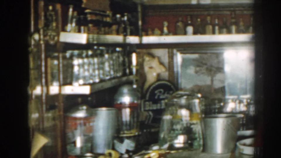 1963 New Jersey home-movie frame showing an indoor bar shelf with glassware, liquor bottles, metal drinkware, and a Pabst sign.