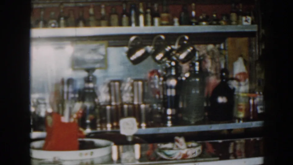 1963 New Jersey indoor bar view showing lower shelves, small lamps, metal drinkware, bottles, glasses, and red bar utensils.