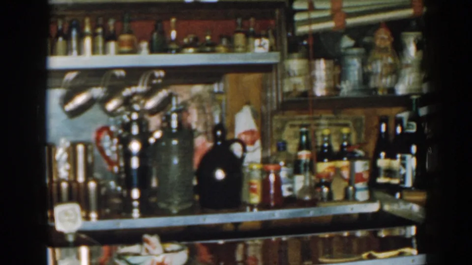 1963 New Jersey home-movie still of a bar counter area with lamps, stocked shelves, metal drinkware, jars, and condiment bottles.