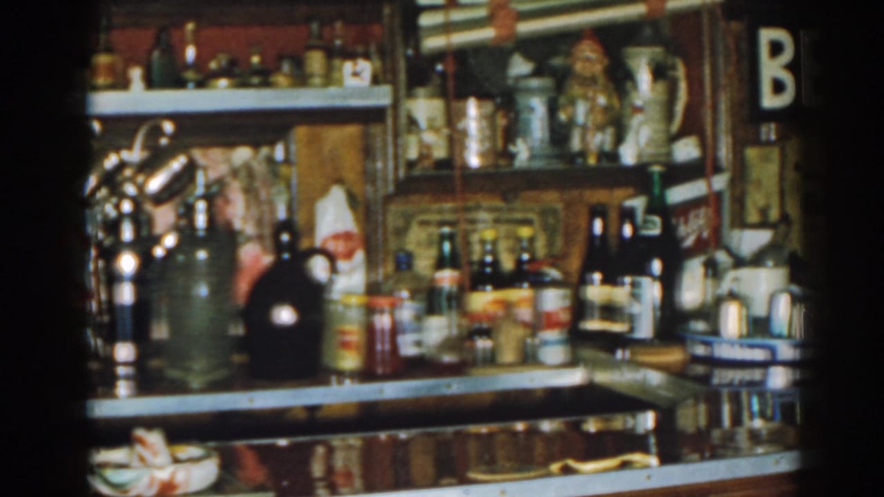 Back bar with bottles, mugs, counter clutter, and shelves in a dim tavern interior.