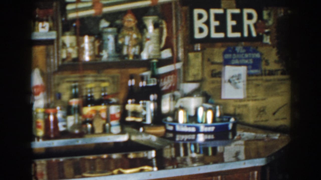 1963 New Jersey tavern interior with bar bottles, mugs, coaster trays, wall notices, and partial BEER signage in soft 8mm focus.