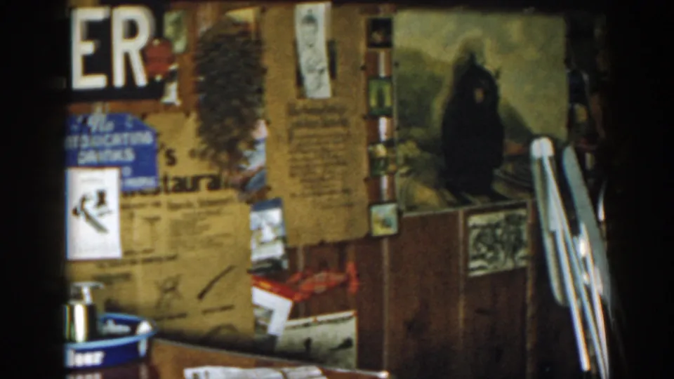 1963 New Jersey home-movie frame of a tavern wall with partial BEER sign, posted items, bottles, counter, and a dark painted figure.
