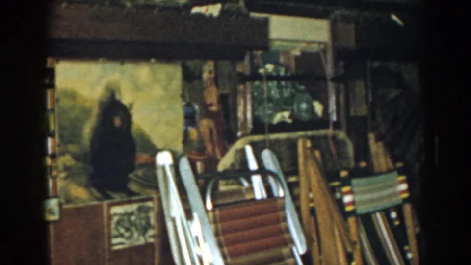 1963 New Jersey 8mm frame with stacked folding chairs, a dark wall painting, wooden boards, and stored items in a private interior.