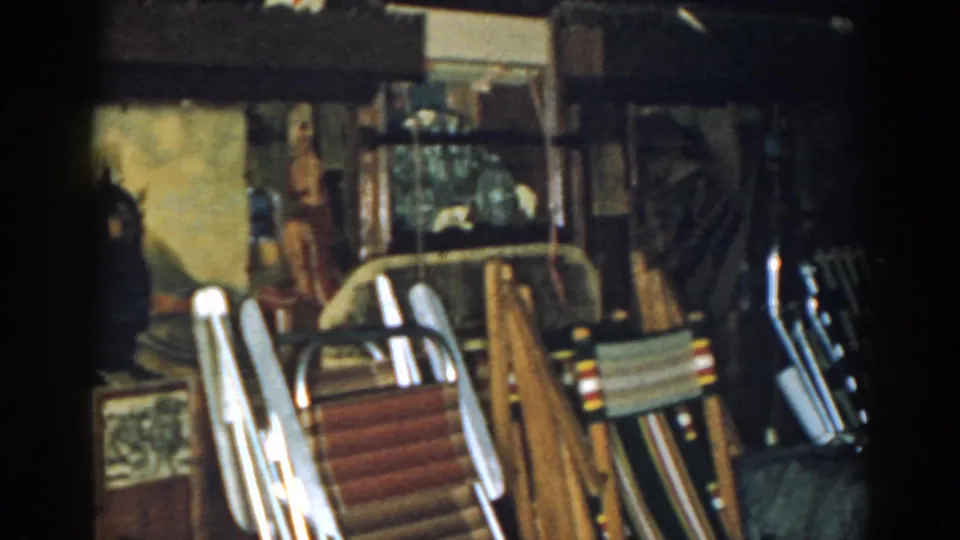 1963 New Jersey home-movie frame showing a cluttered storage interior with folding chairs, boards, papers, a wall painting, and glassed area.