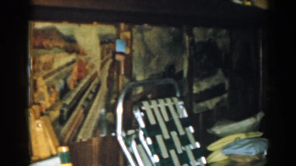 1963 New Jersey basement frame with a train mural, metal folding chair, stacked items, and soft 8mm color shifts.
