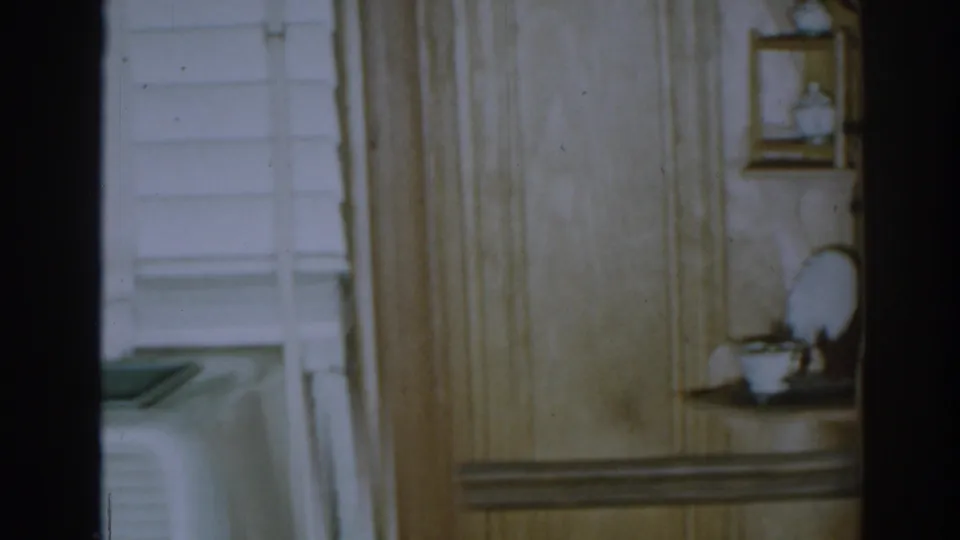 1963 New Jersey home-movie frame with wood paneling, a pale window blind, and a small shelf displaying white ceramic dishes.