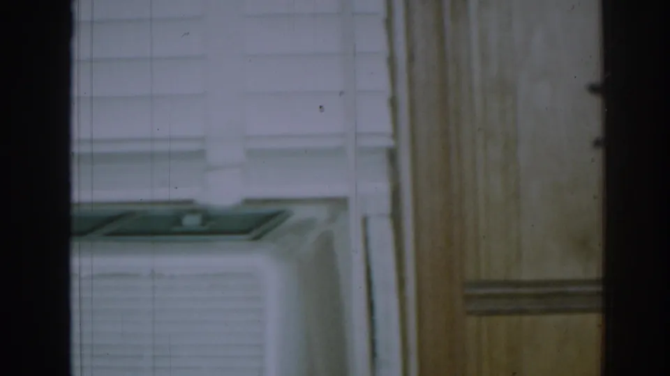 1963 New Jersey home-movie frame showing a white window-mounted air conditioner behind dark vertical framing and wood trim.