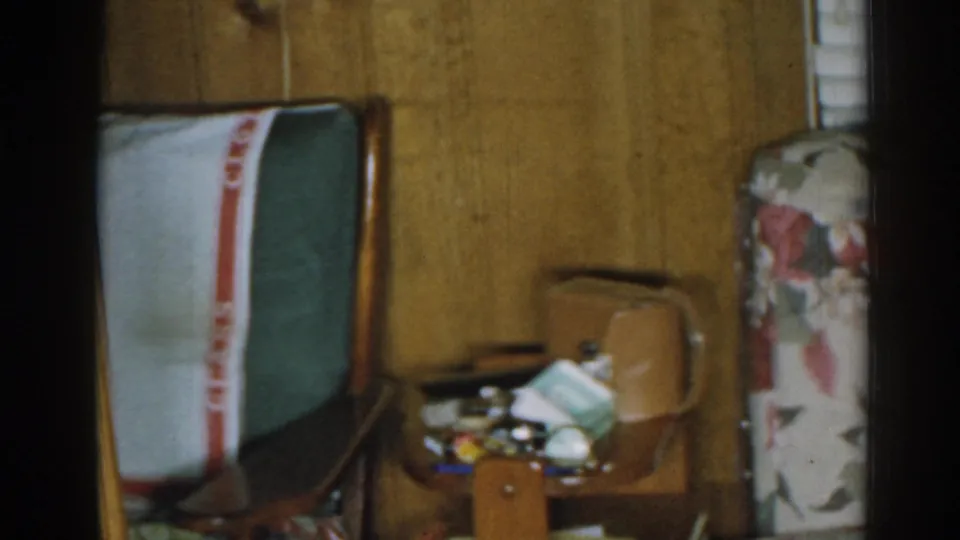 1963 New Jersey home-movie frame showing a wood-paneled living room with floral seating, a side table, and a wooden chair.